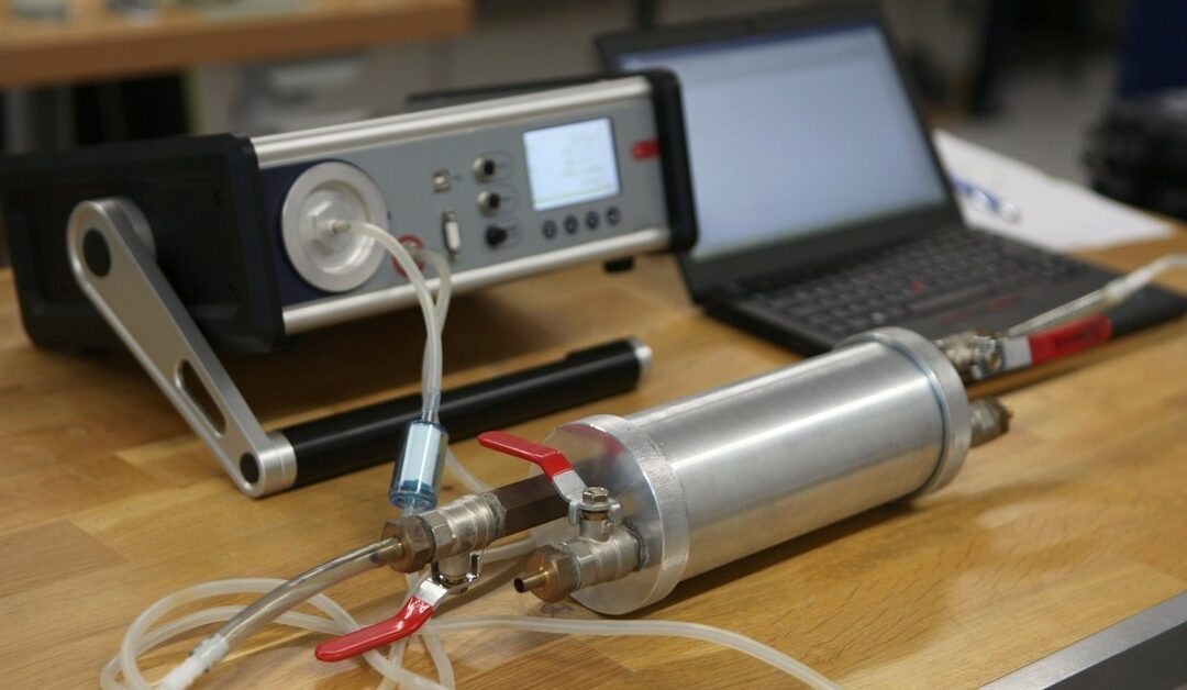 A traditional radon gas detector next to a laptop on a table. There's a piece of paper next to the laptop with a pen.