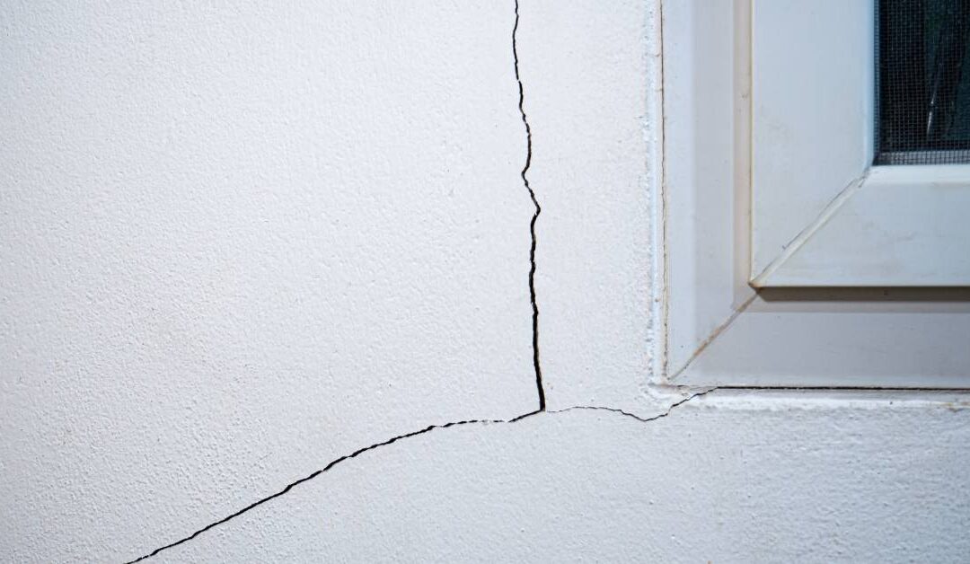 A crack on a wall with a window next to it. The window has a screen behind it and the wall is white.