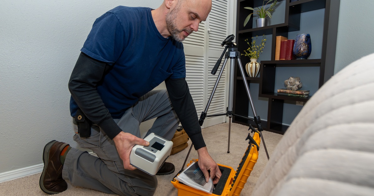 A man wearing a blue shirt and gray pants kneels in a room as he is sets up radon testing equipment.