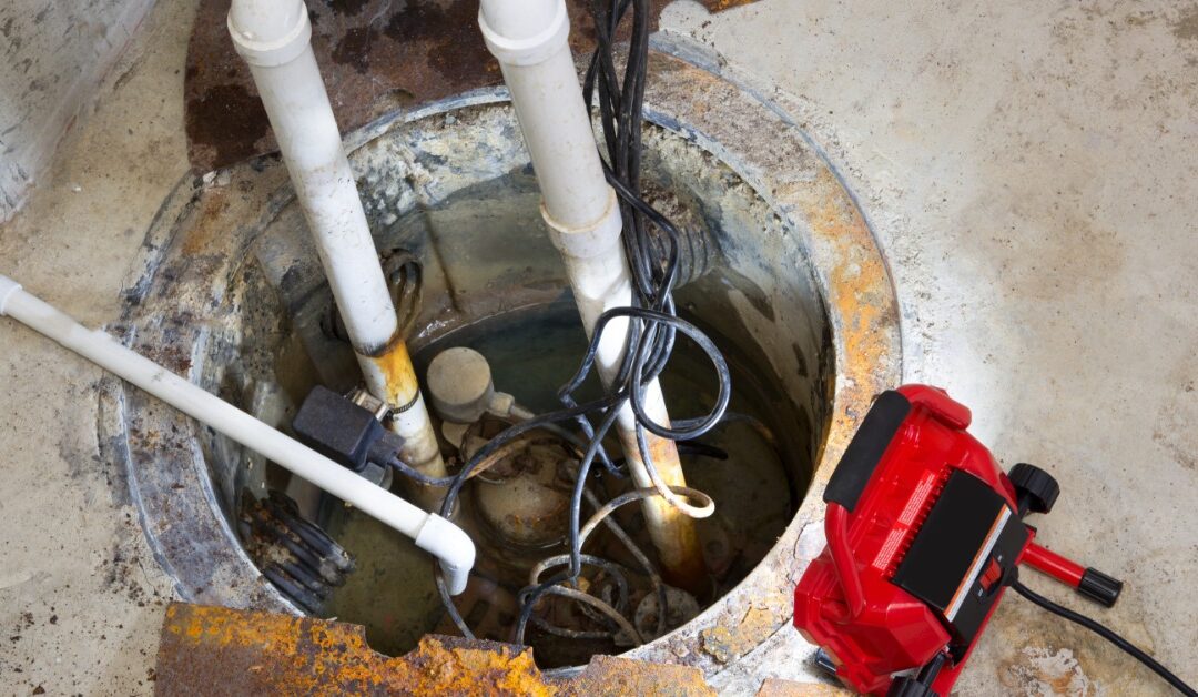 An open sump pit holds water, pipes, and pump equipment as a red work light rests on the concrete basement floor.